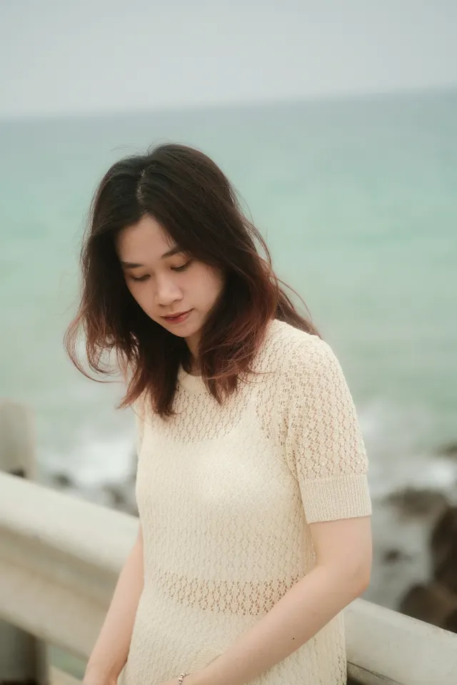 A woman with long dark hair looking down, the sea soft behind her
