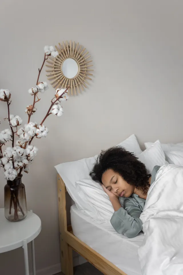 A woman asleep in white bedding beside a vase of cotton stems